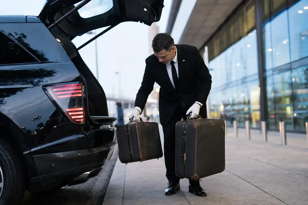 Chauffeur in white gloves lifting luggage onto the curb beside a black luxury SUV at airport departures